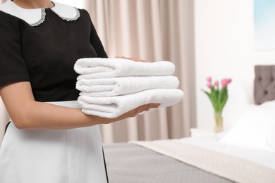Young maid holding stack of fresh towels in hotel room, closeup