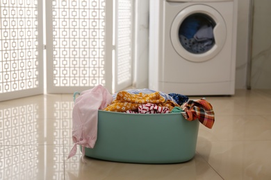 Photo of Plastic laundry basket full of different clothes on floor in bathroom