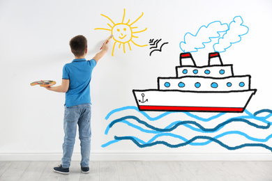 Image of Cute child drawing sea with cruise ship on white wall indoors