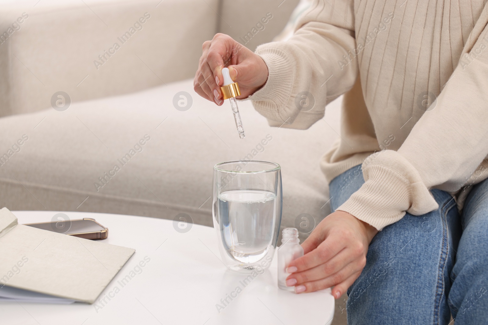 Photo of Woman dripping food supplement into glass of water on white table indoors, closeup