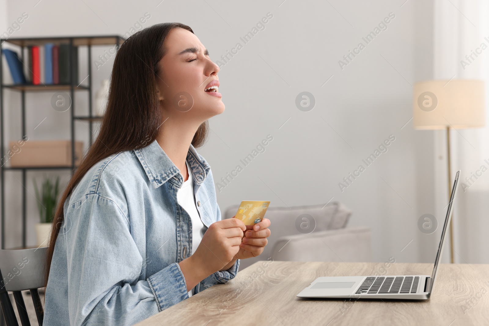 Photo of Upset woman with credit card and laptop at table indoors. Be careful - fraud