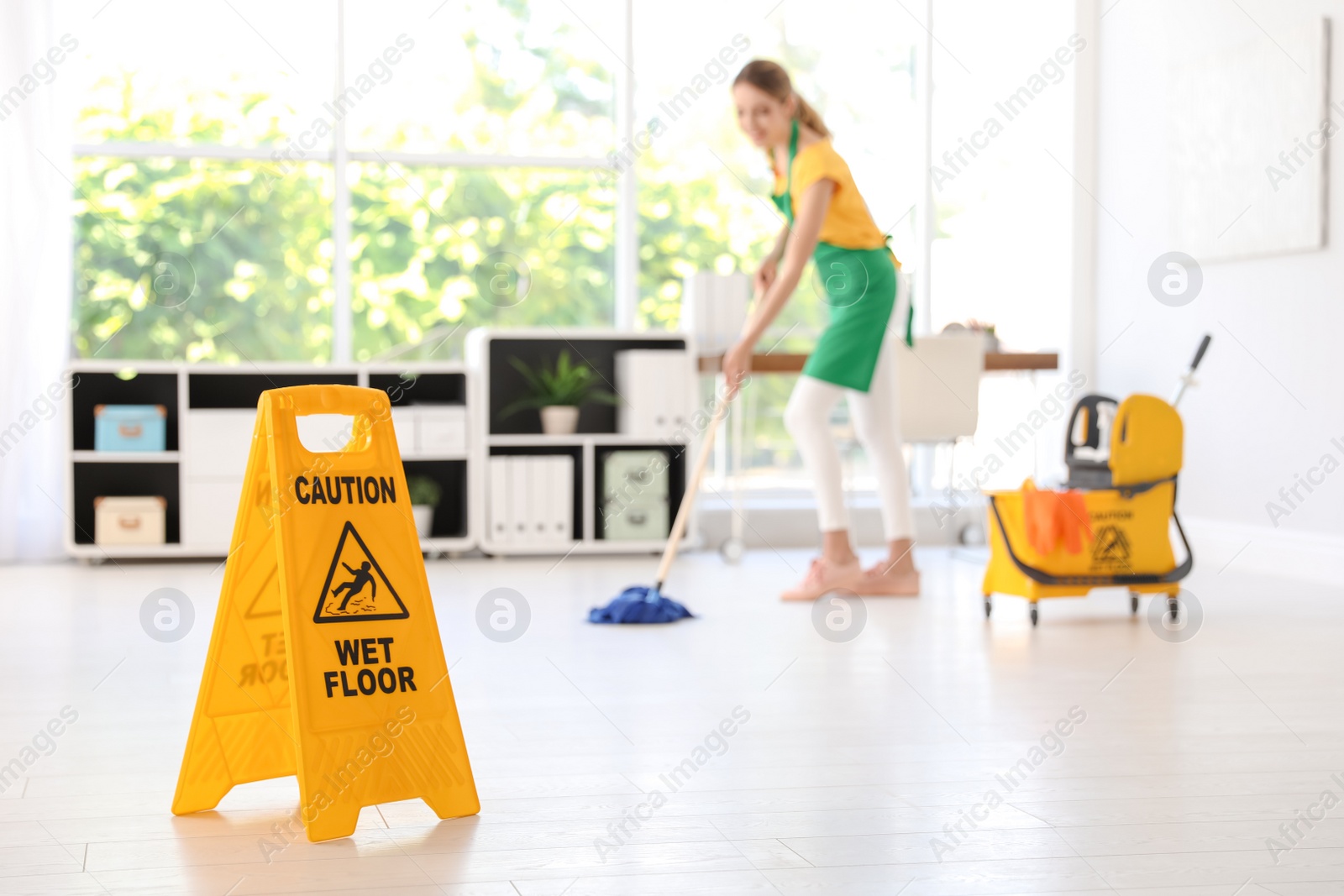 Photo of Safety sign with phrase "CAUTION WET FLOOR" and young woman cleaning in office