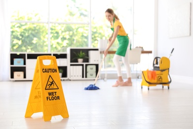 Safety sign with phrase "CAUTION WET FLOOR" and young woman cleaning in office