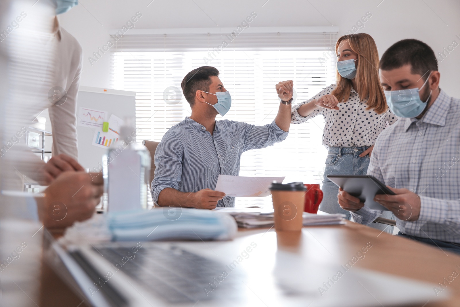 Photo of Coworkers with protective masks making elbow bump in office. Informal greeting during COVID-19 pandemic