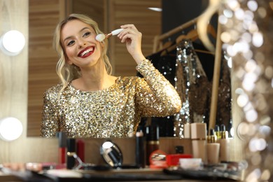 Photo of Beautiful makeup. Smiling woman applying powder with brush onto face in front of mirror in dressing room