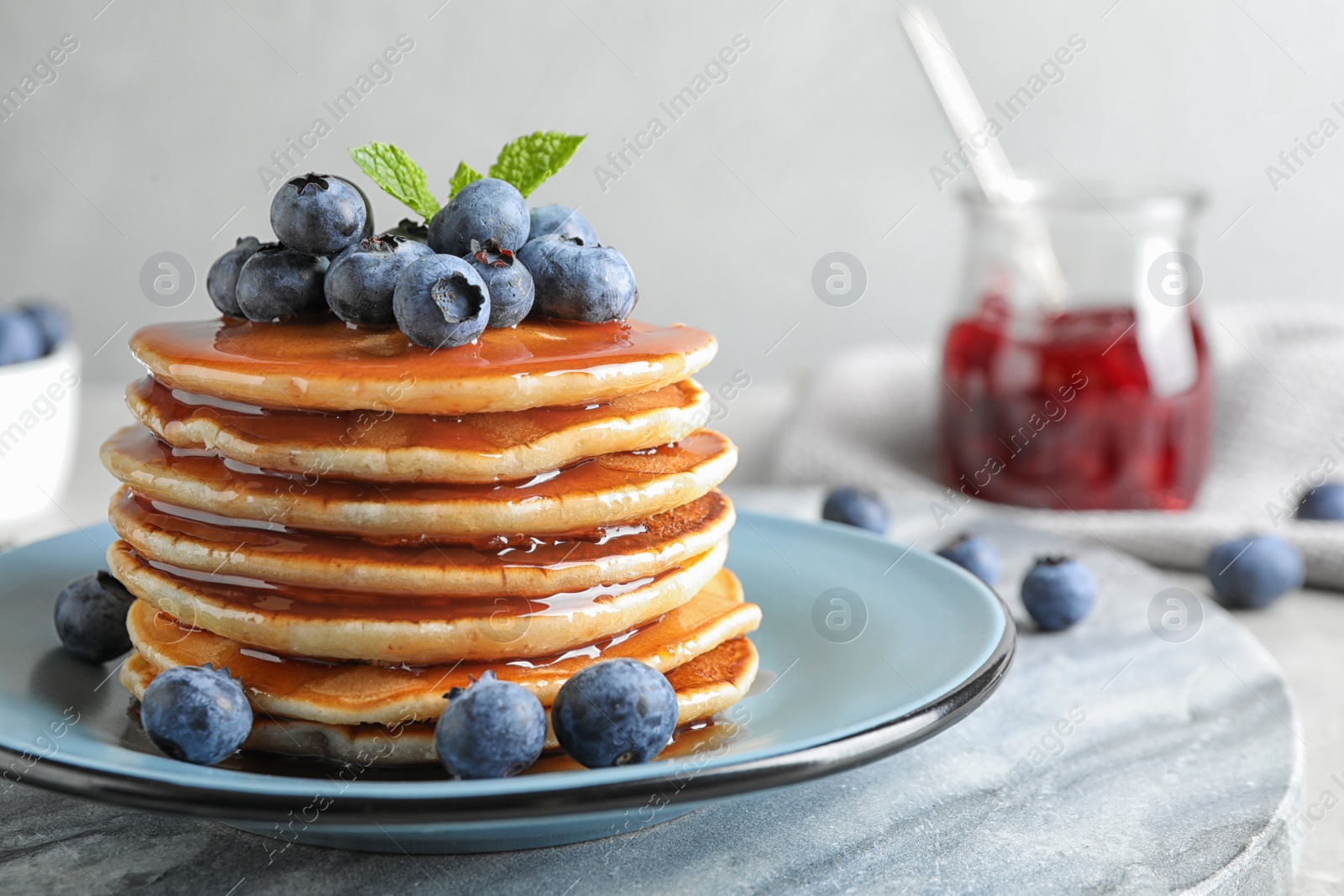 Photo of Plate of delicious pancakes with fresh blueberries and syrup on grey table against light background