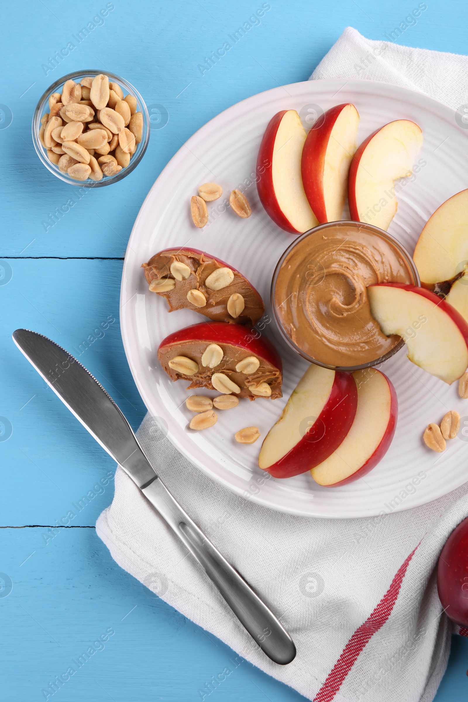 Photo of Slices of fresh apple with peanut butter, nuts and knife on light blue wooden table, flat lay