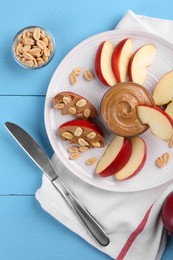 Slices of fresh apple with peanut butter, nuts and knife on light blue wooden table, flat lay