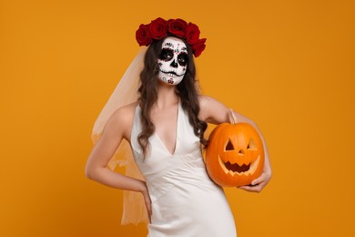 Photo of Young woman in scary bride costume with sugar skull makeup, flower crown and carved pumpkin on orange background. Halloween celebration