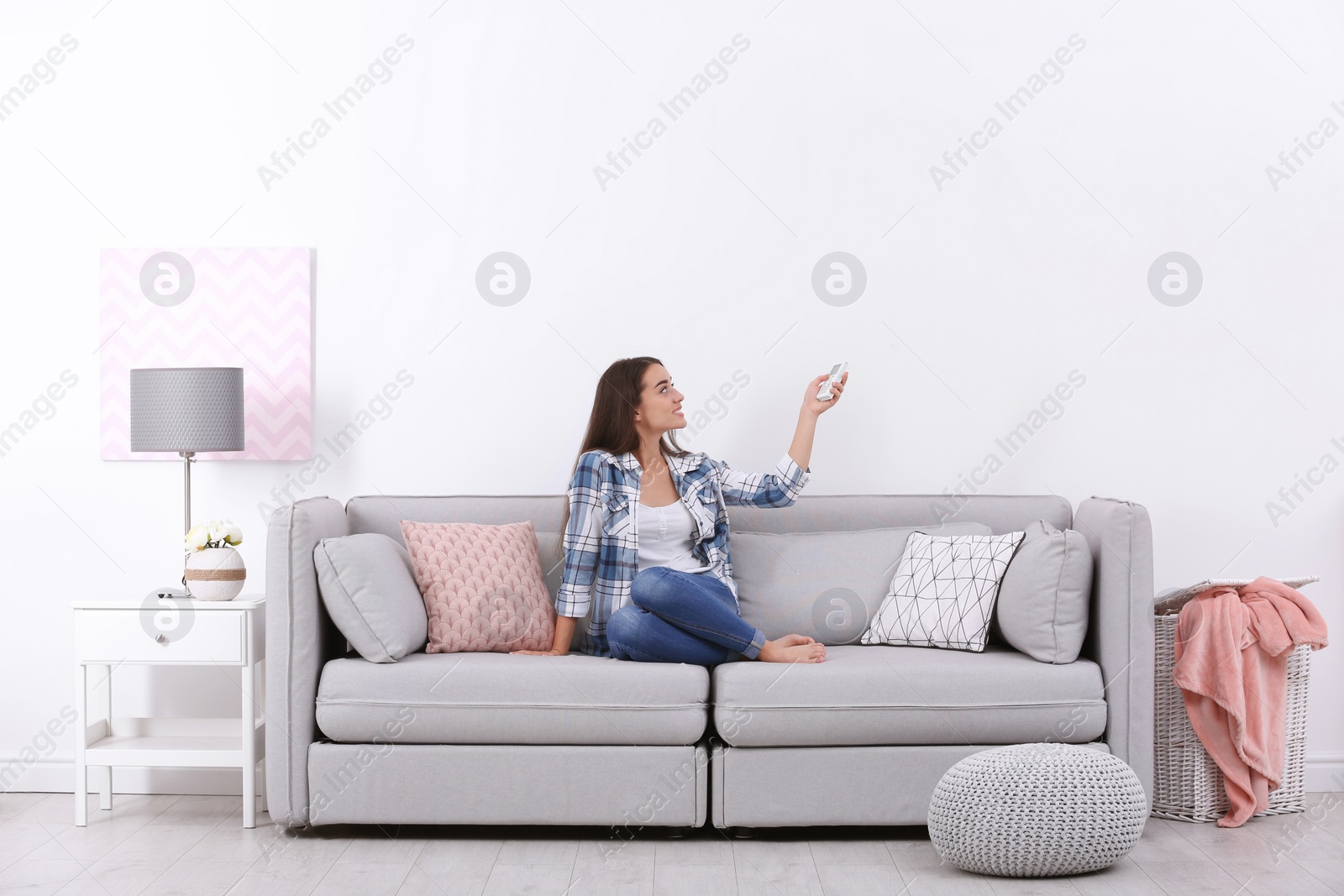 Photo of Young woman switching on air conditioner while sitting on sofa near white wall