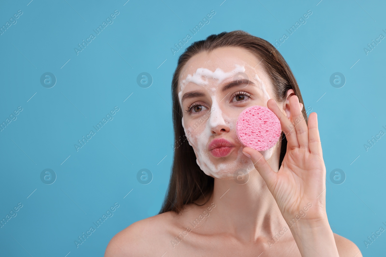 Photo of Happy young woman washing her face with sponge on light blue background. Space for text