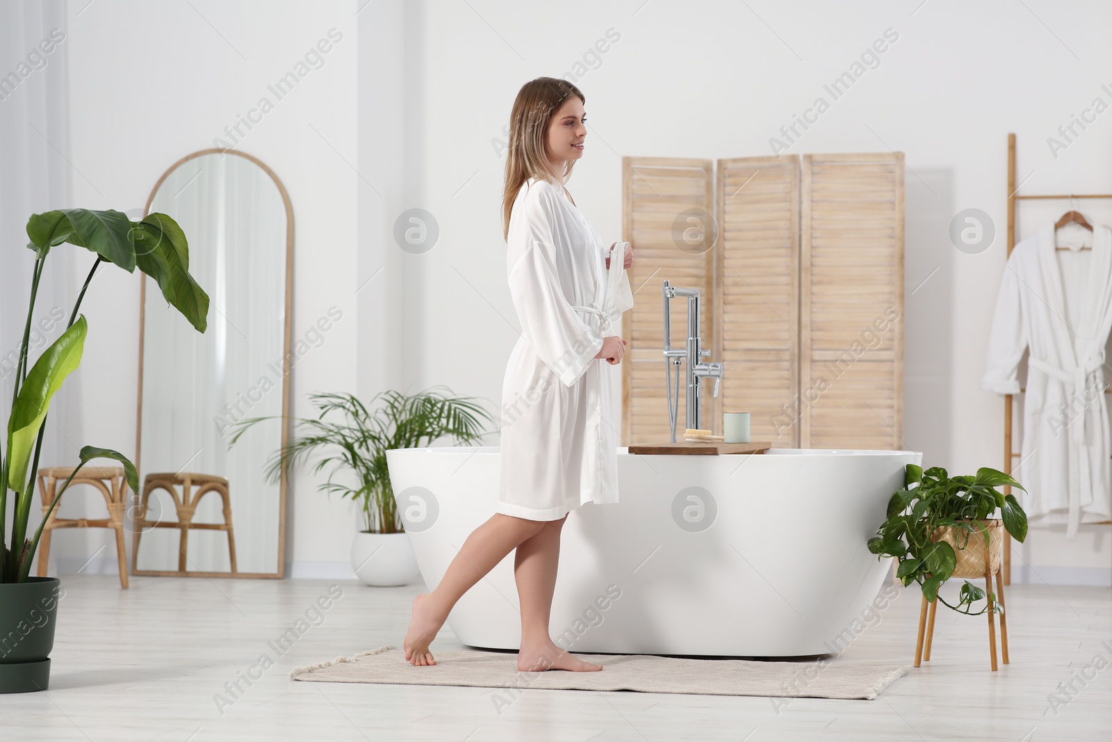 Photo of Beautiful happy woman wearing white robe near tub in bathroom