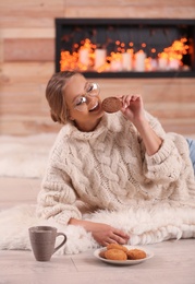 Photo of Beautiful young woman eating cookie on rug at home. Winter atmosphere
