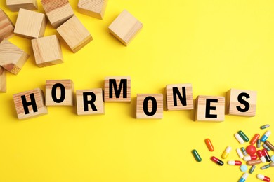 Word Hormones made of wooden cubes with letters and pills on yellow background, flat lay