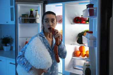 Photo of Young woman with pillow eating sausages near open refrigerator at night