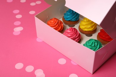 Box with different cupcakes and confetti on pink background, space for text