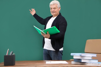 Portrait of senior teacher with notebook at green chalkboard and table