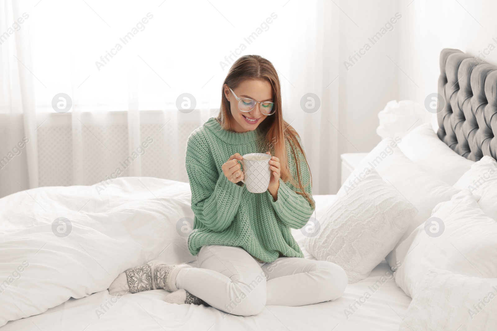 Photo of Young woman in warm sweater with hot drink on bed at home