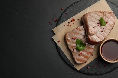 Photo of Delicious tuna steaks with sauce, parsley and spices on black table, top view. Space for text