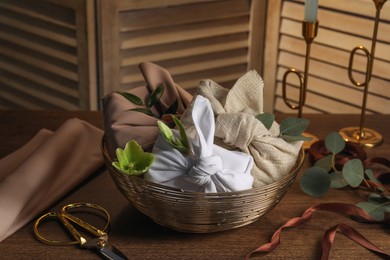 Photo of Furoshiki technique. Gifts packed in different fabrics, flowers, leaves, scissors and ribbon on wooden table