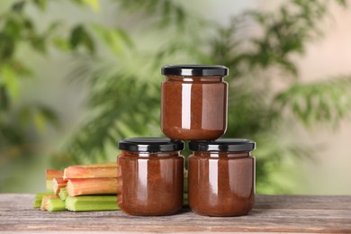 Photo of Jars of tasty rhubarb jam and stalks on wooden table, closeup