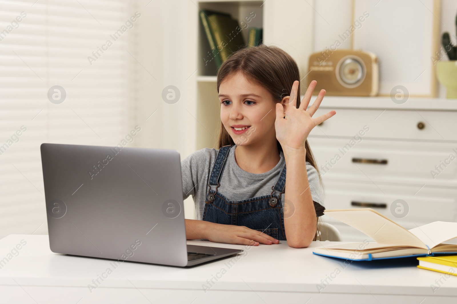 Photo of Cute girl waving hello during online lesson via laptop at white table indoors