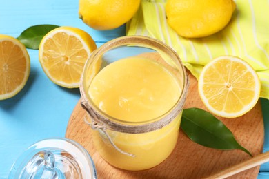 Delicious lemon curd in glass jar, fresh citrus fruits and green leaves on light blue wooden table, closeup
