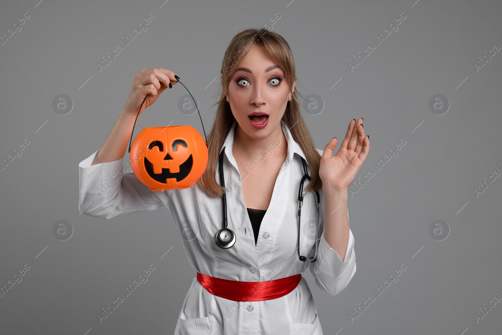 Photo of Emotional woman in scary nurse costume with pumpkin bucket on light grey background. Halloween celebration