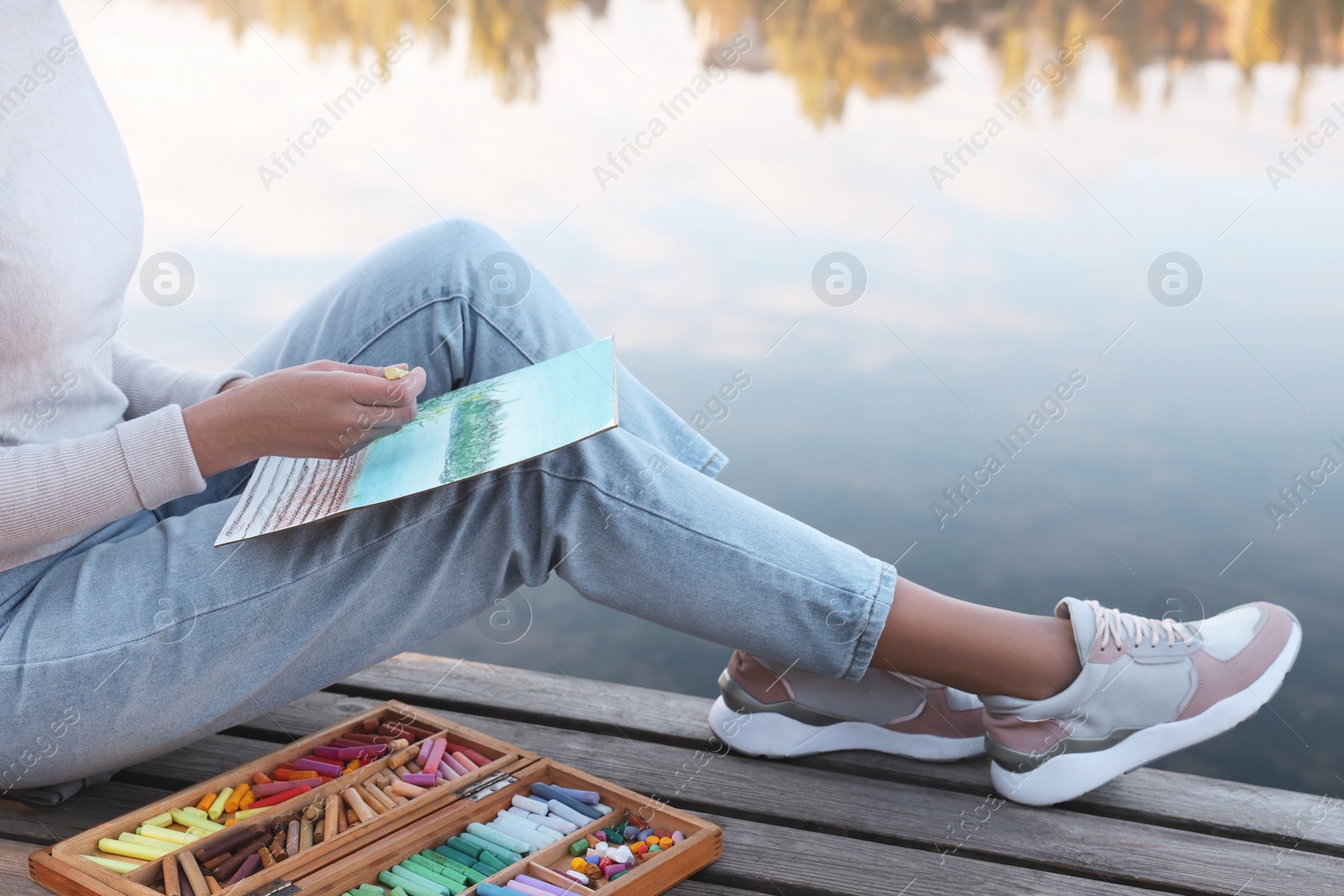 Photo of Woman drawing with soft pastels on wooden pier near river, closeup