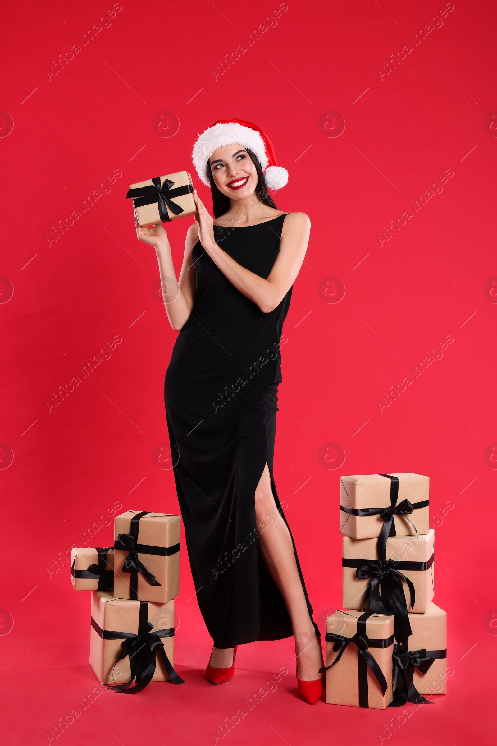 Photo of Woman in black dress and Santa hat with Christmas gifts on red background