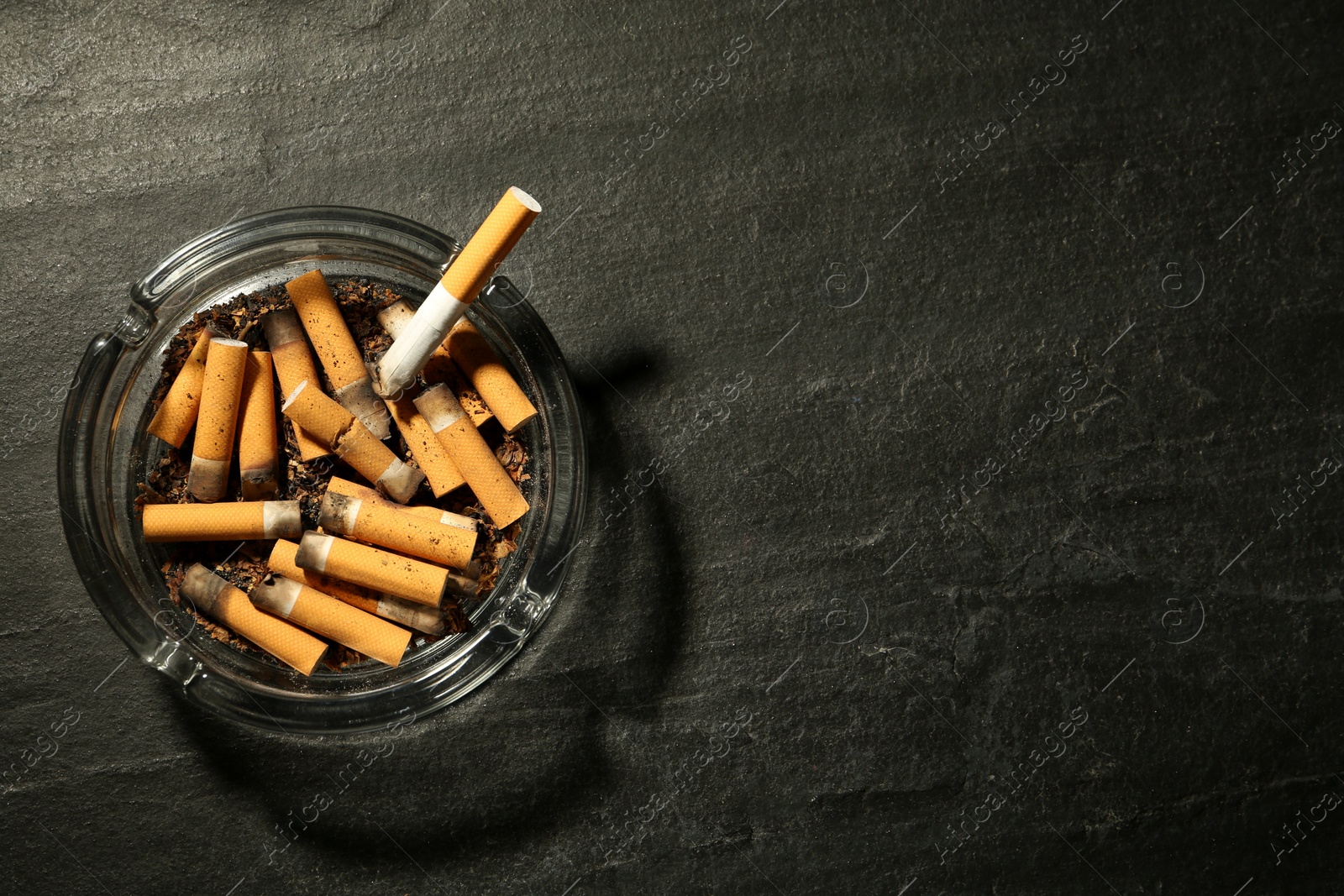 Photo of Glass ashtray with cigarette stubs on dark grey table, top view. Space for text