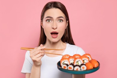 Emotional young woman with plate of sushi rolls and chopsticks on pink background