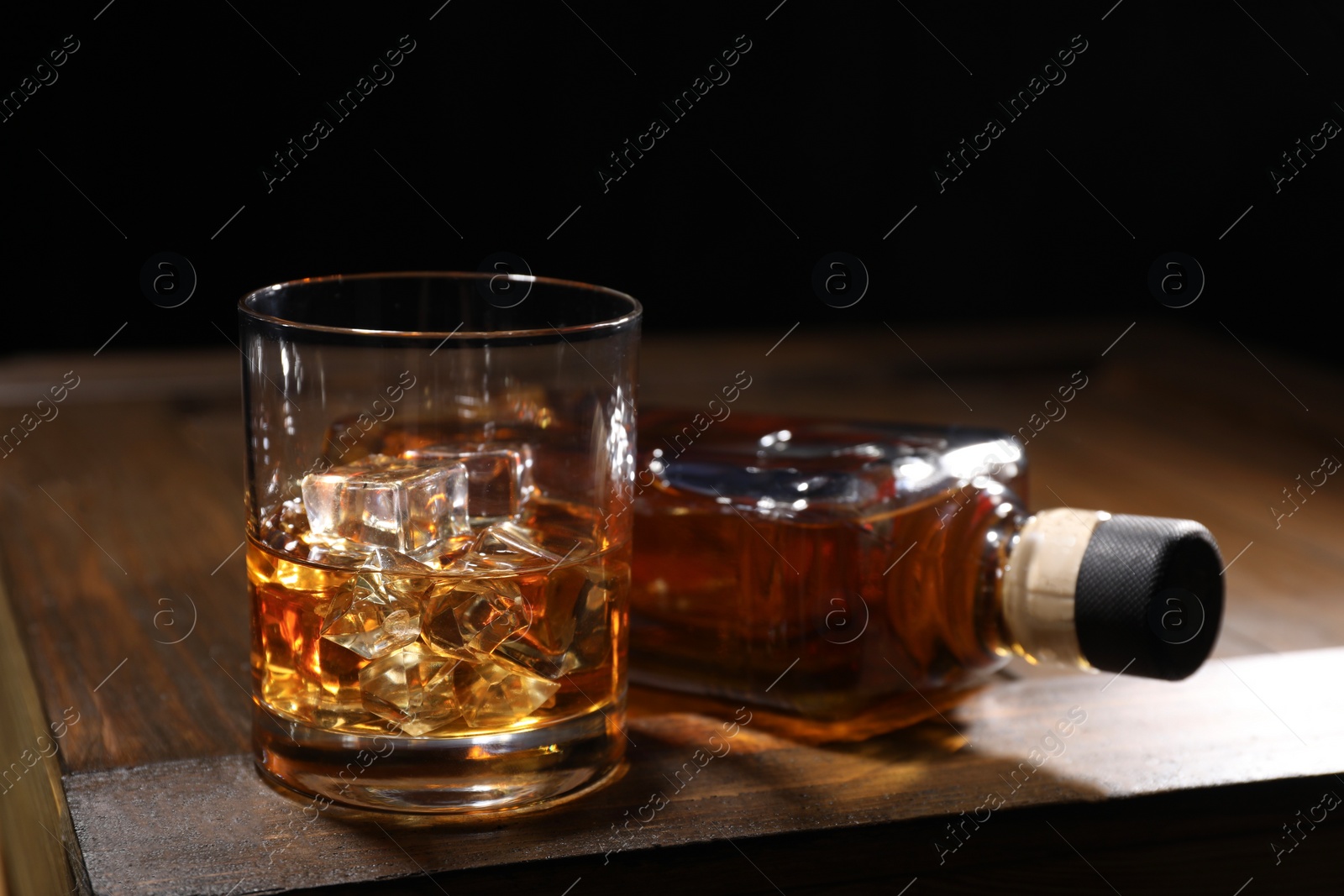 Photo of Whiskey with ice cubes in glass and bottle on wooden crate against black background, closeup