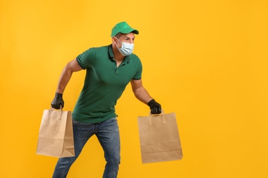 Photo of Courier in medical mask holding paper bags with takeaway food on yellow background, space for text. Delivery service during quarantine due to Covid-19 outbreak