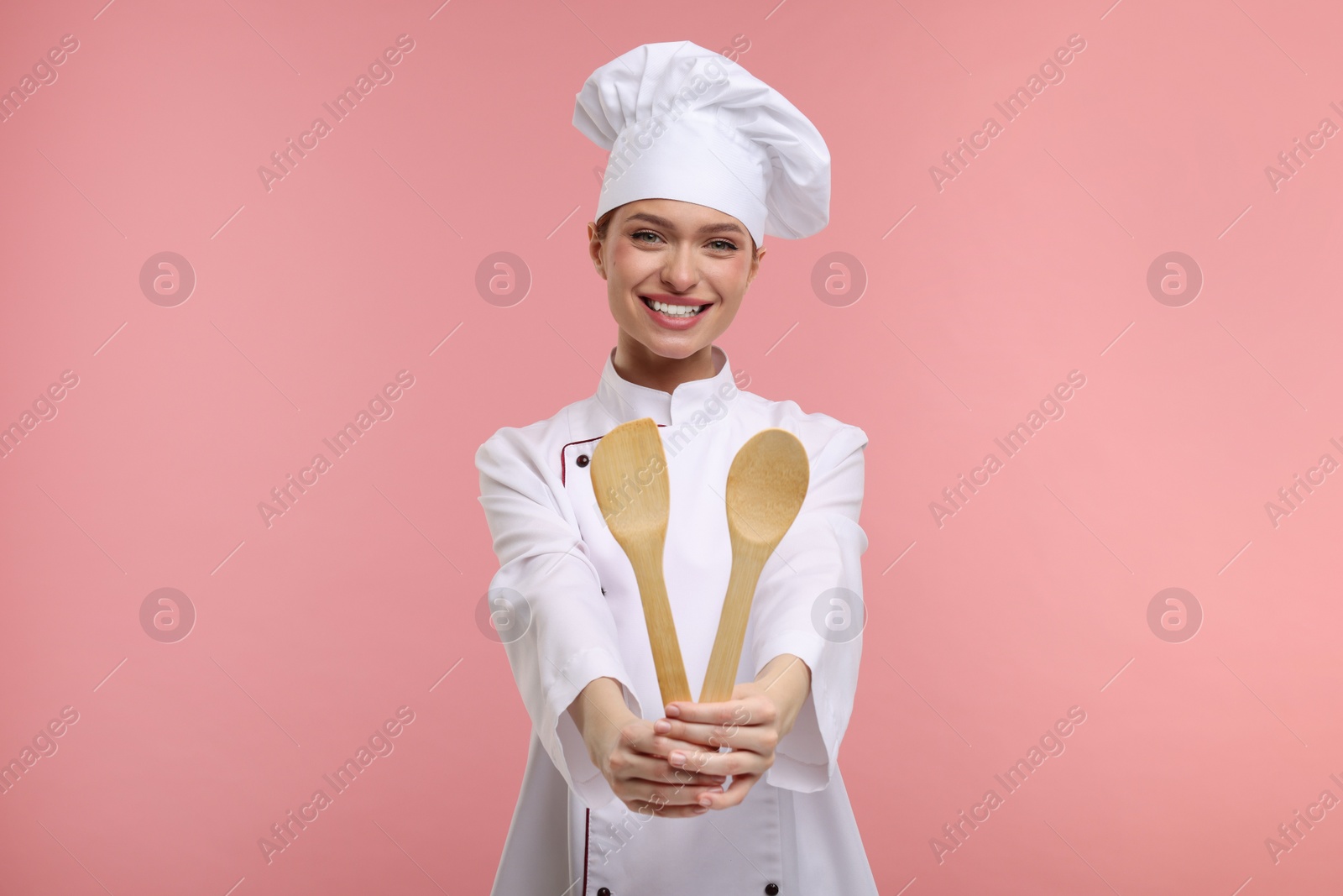 Photo of Happy chef in uniform holding wooden spatula and spoon on pink background