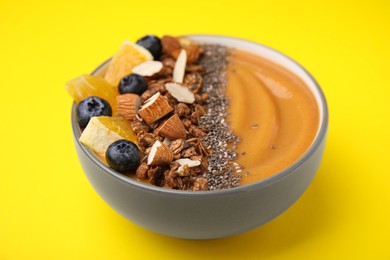 Photo of Bowl of delicious fruit smoothie with fresh orange slices, blueberries and granola on yellow background, closeup
