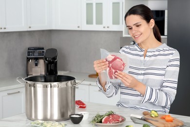Photo of Woman holding vacuum packed meat near pot with sous vide cooker at table in kitchen. Thermal immersion circulator