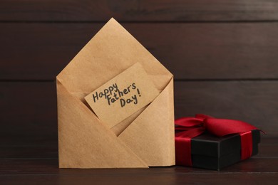 Photo of Card with phrase Happy Father's Day in envelope and gift box on wooden table, closeup