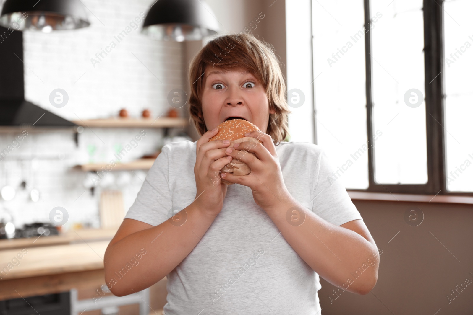 Photo of Emotional overweight boy with tasty burger in kitchen