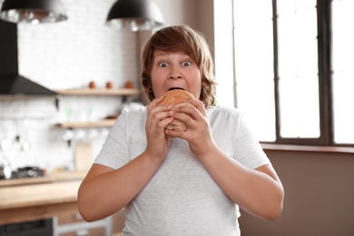 Emotional overweight boy with tasty burger in kitchen