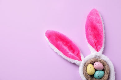 Photo of Headband with Easter bunny ears and dyed eggs in nest on violet background, flat lay. Space for text