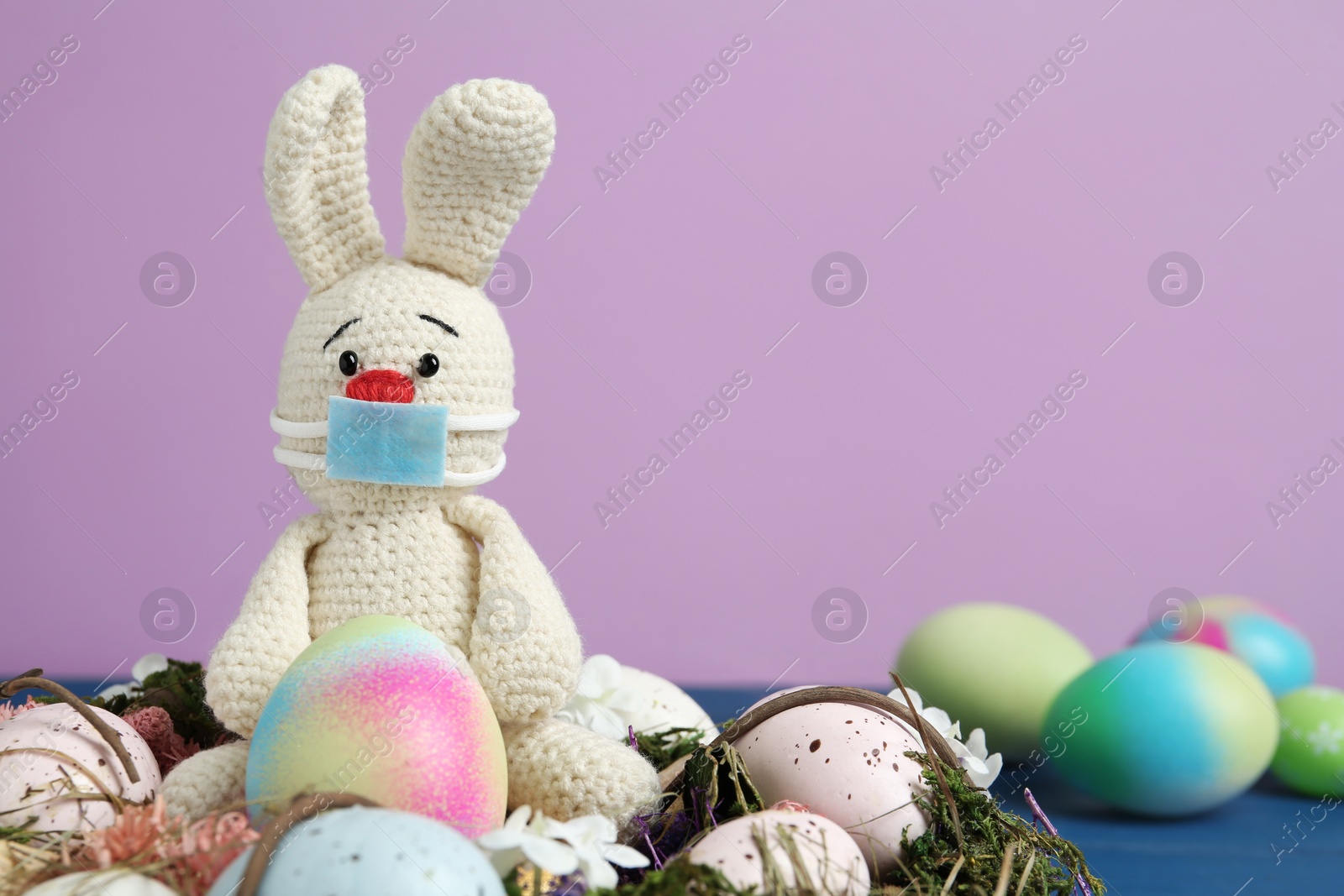 Photo of Toy bunny with protective mask and painted eggs on blue wooden table, space for text. Easter holiday during COVID-19 quarantine