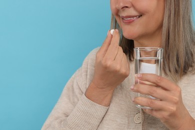 Senior woman with glass of water taking pill on light blue background, closeup