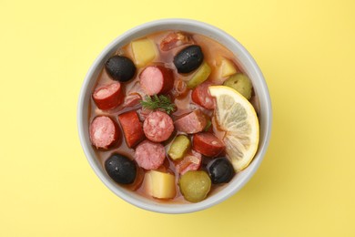 Meat solyanka soup with thin dry smoked sausages in bowl on pale yellow background, top view