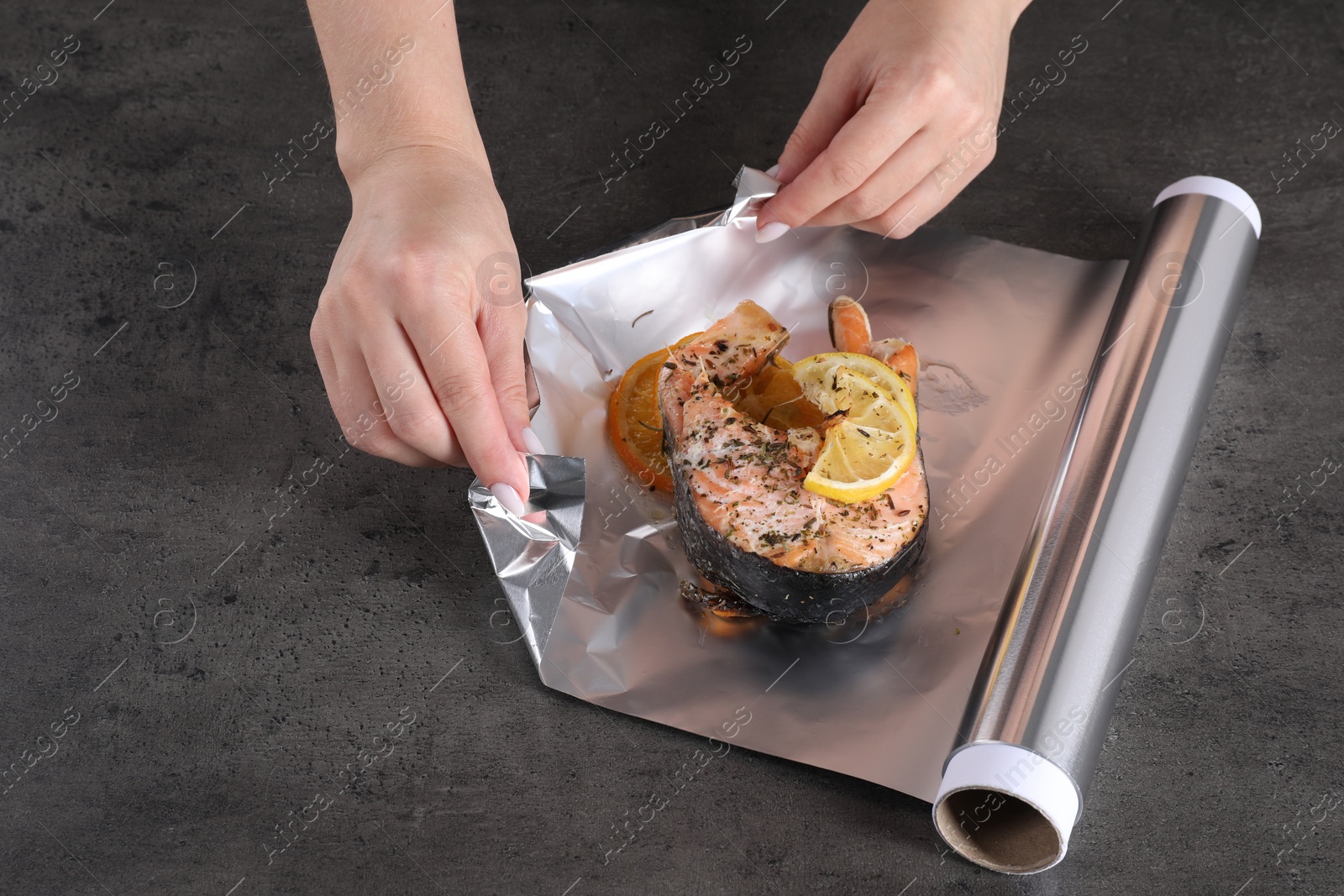 Photo of Woman wrapping tasty salmon with lemon and spices in aluminum foil at grey textured table, closeup