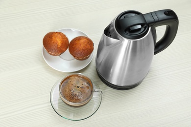 Modern electric kettle, cup of coffee and muffins on white wooden table