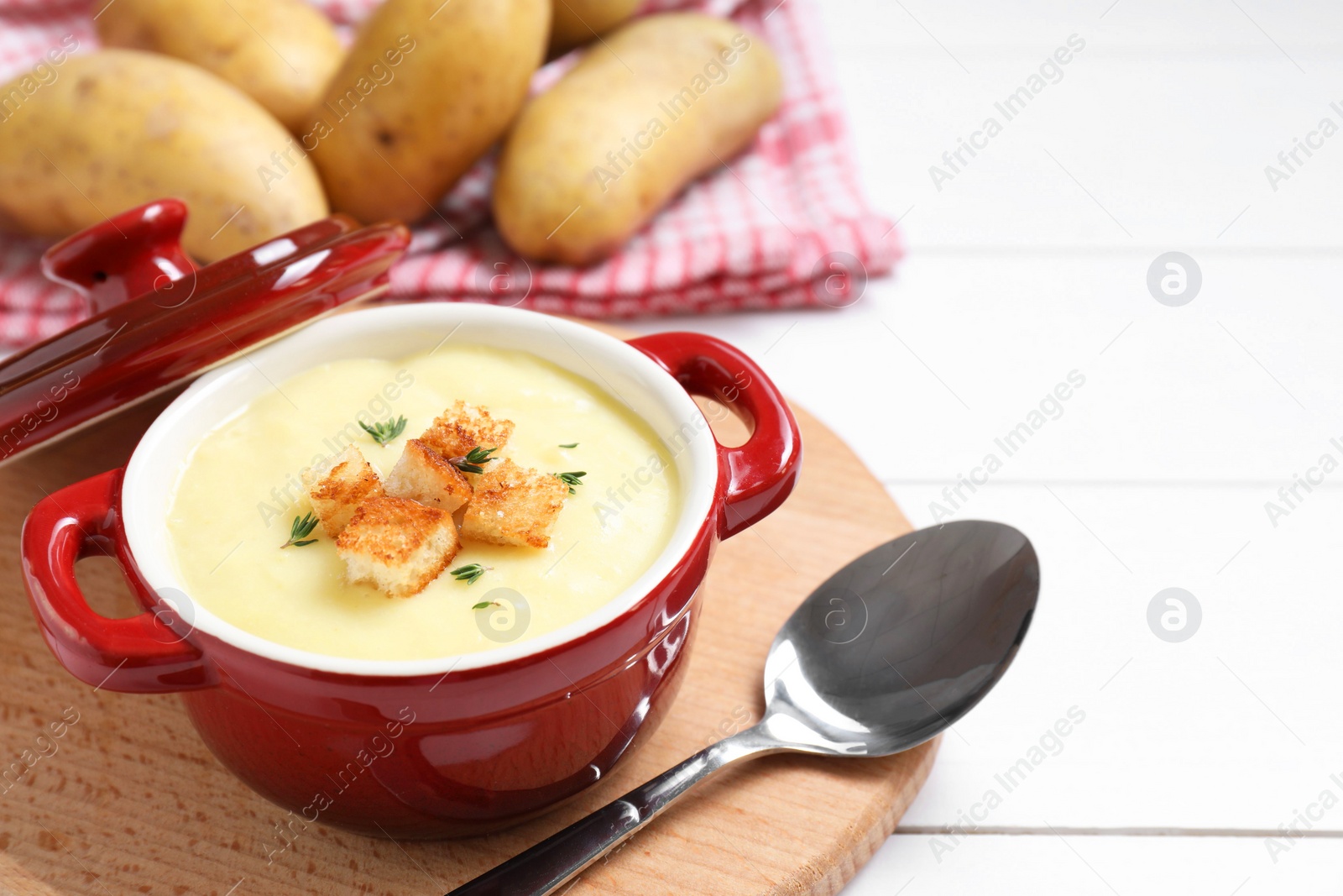 Photo of Tasty potato soup with croutons in ceramic pot and spoon on white wooden table