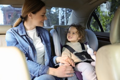 Photo of Cute little girl with toy rabbit sitting in child safety seat near mother inside car