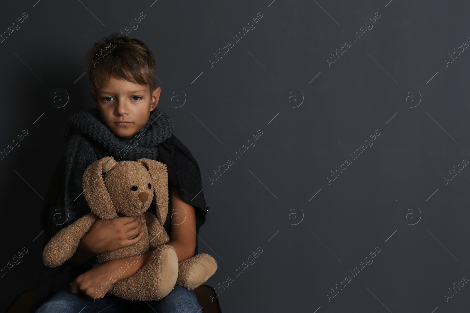 Photo of Poor boy sitting with toy rabbit on dark background. Space for text