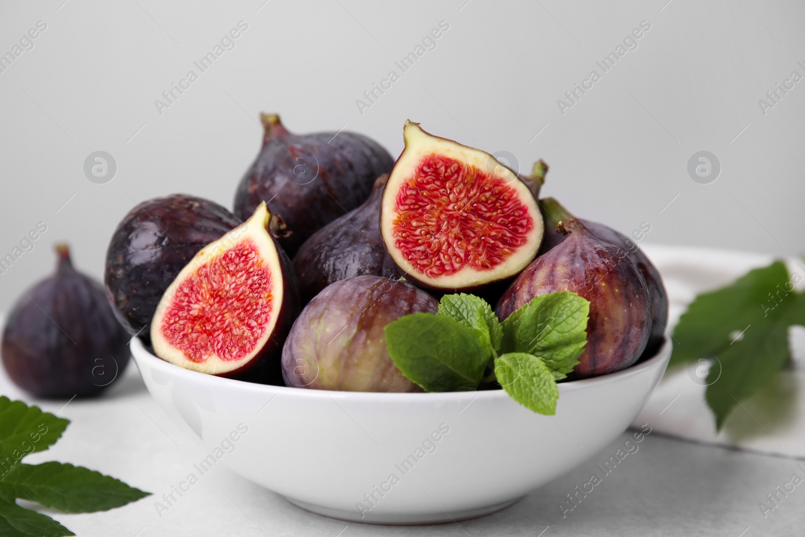 Photo of Bowl of tasty ripe figs and green leaves on light table, closeup